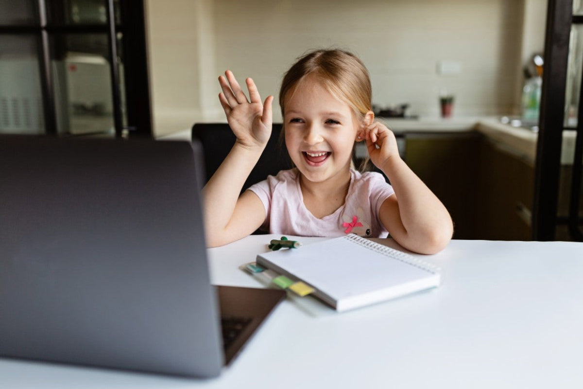 Smiling young girl waving at a laptop while sitting at a table with a notebook, representing the value of refurbished laptops for learning