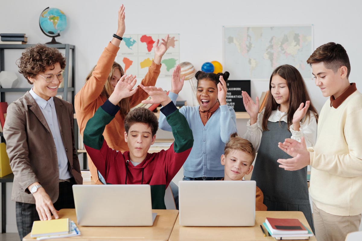 Group of children and a teacher in a classroom setting with laptops and raised hands