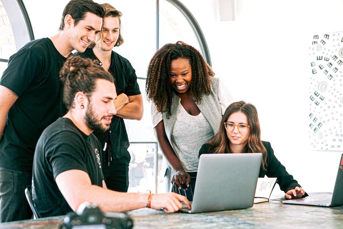 Group of young professionals gathered around a laptop, discussing refurbished electronics in a modern office setting