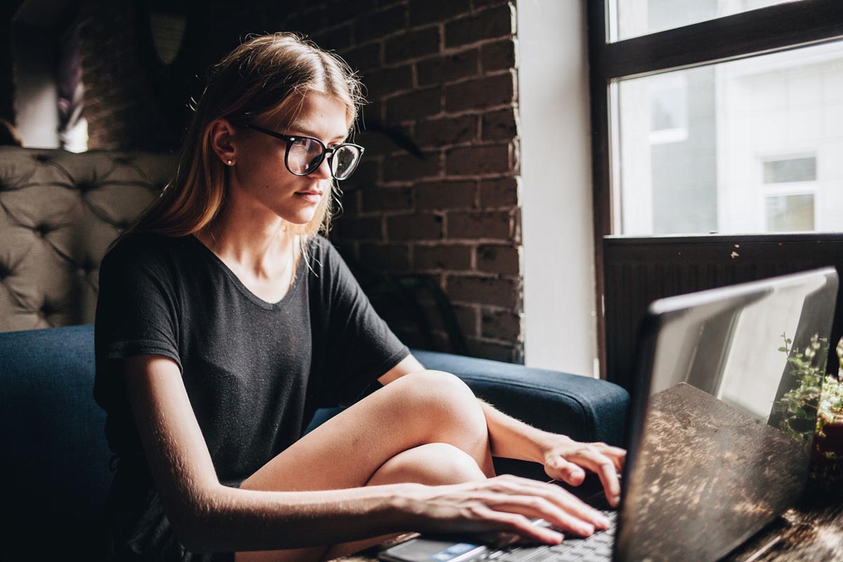 Young woman using a laptop on a couch near a window, illustrating the practical use of refurbished laptops